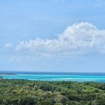 A panoramic view of a lush green landscape meeting a vibrant turquoise sea under a clear blue sky with scattered clouds.
