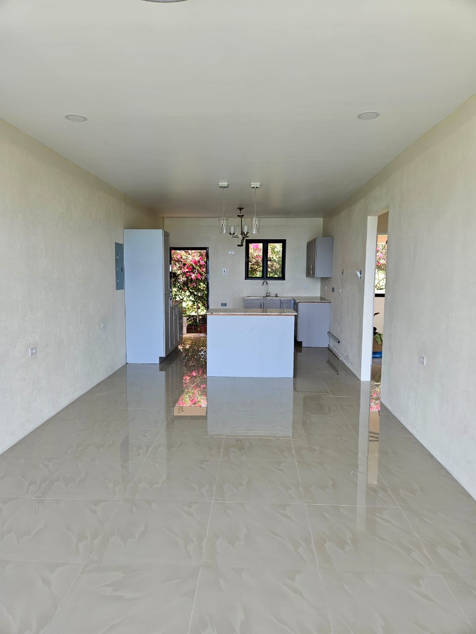 Interior view of a spacious, modern kitchen and living area with light-colored walls and glossy tiled floor, featuring a refrigerator, a sink, and large windows that provide natural light and a view of flowering plants outside.